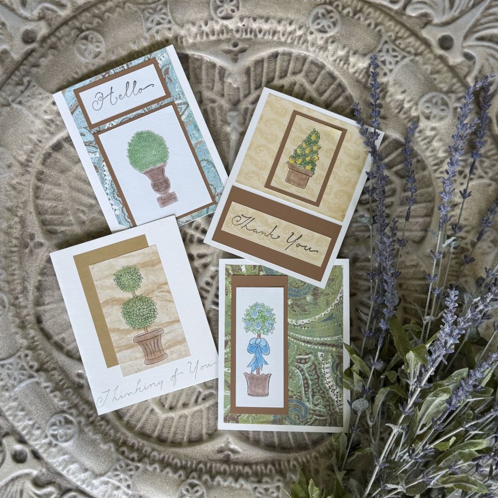 Four greeting cards with potted topiary illustrations and messages like "Hello," "Thank You," and "Thinking of You," are arranged on an ornate textured surface next to a sprig of lavender.
