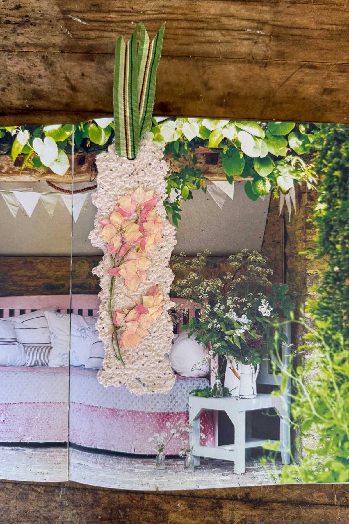 A handmade bookmark with pressed pink flowers and green ribbon rests on an open book showing a cozy, rustic bedroom with a pink bedspread and flowers on a white table. The scene includes greenery and bunting.
