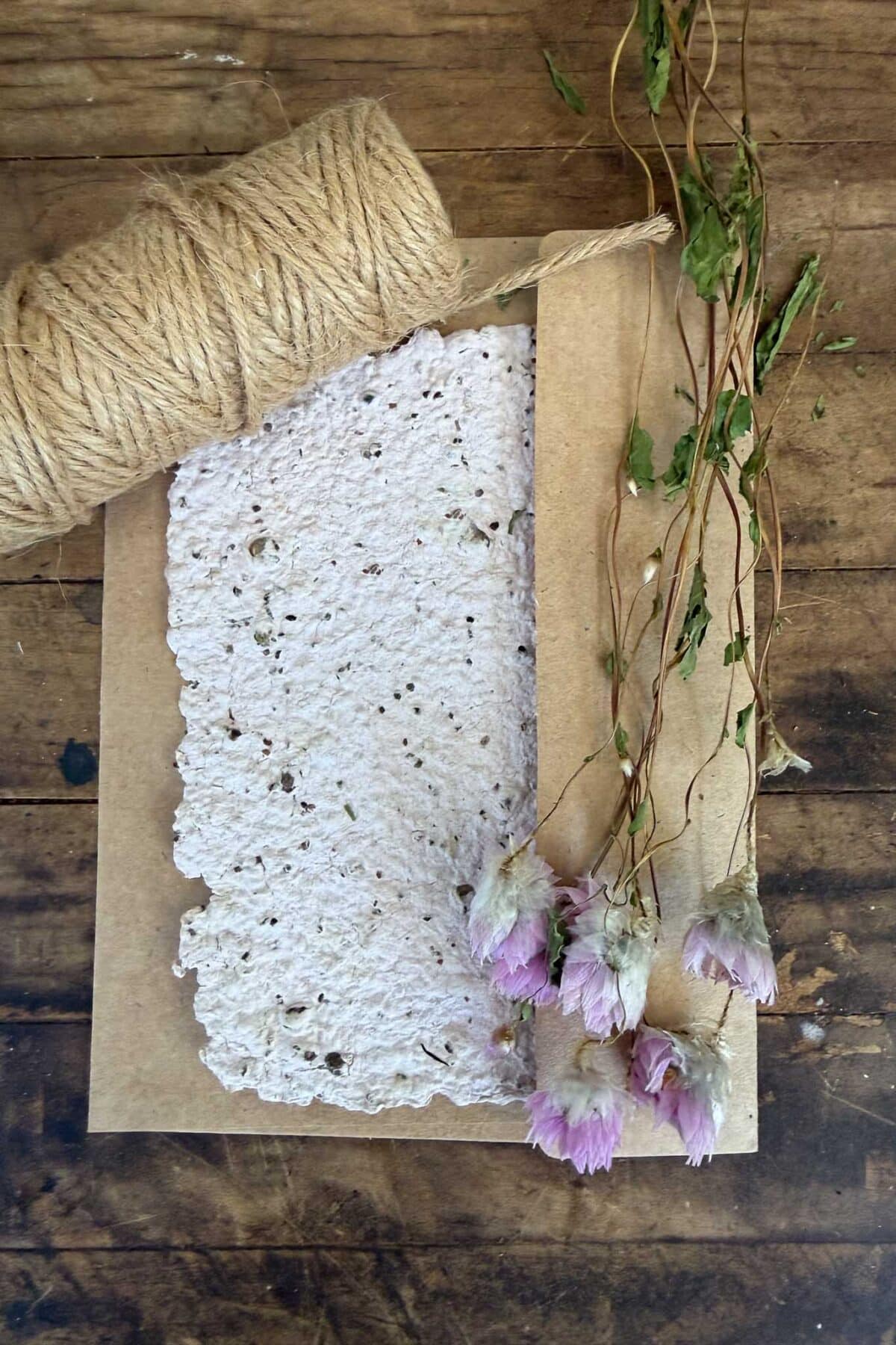 A piece of textured, handmade paper with scalloped edges lies on a brown envelope. Beside it are a spool of twine and several dried pink and white flowers, all on a rustic wooden surface.