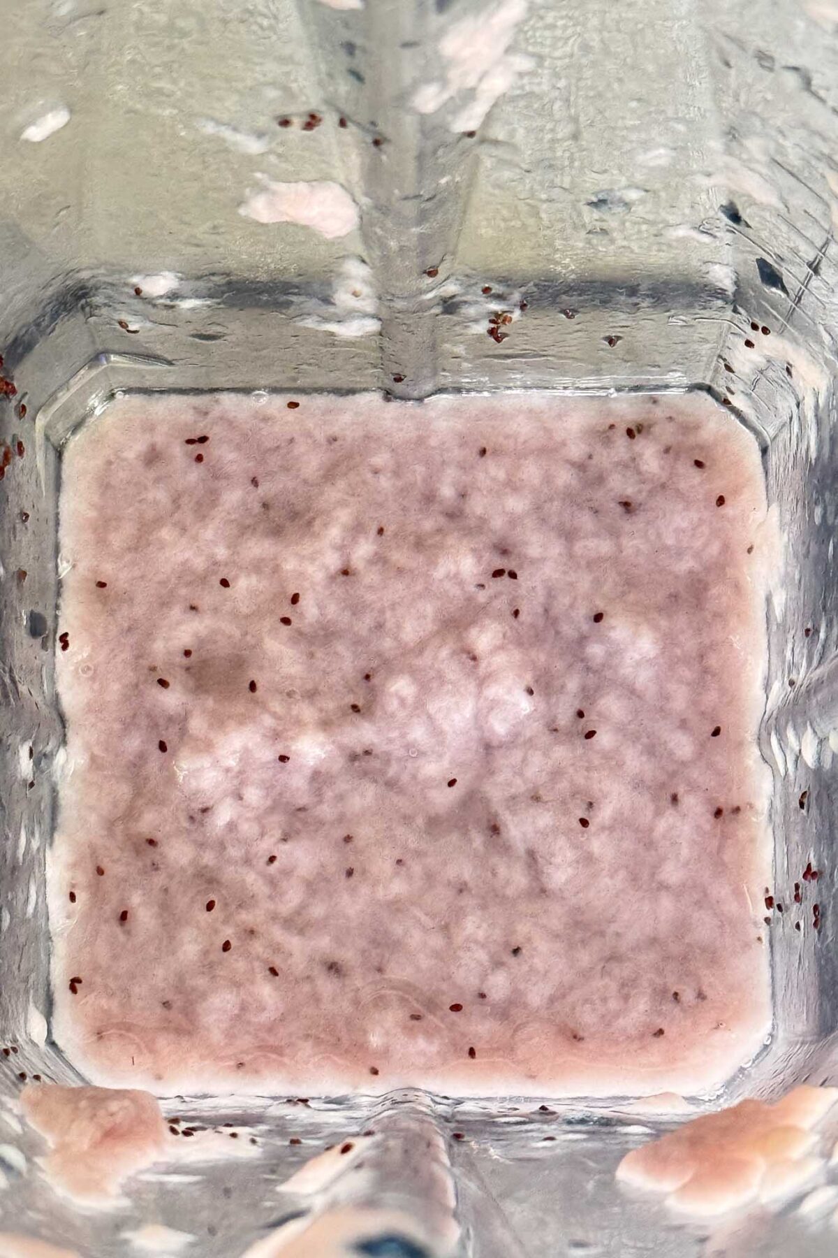 A top-down view of a blender containing a pale pink smoothie mixture with visible small dark seeds scattered throughout.