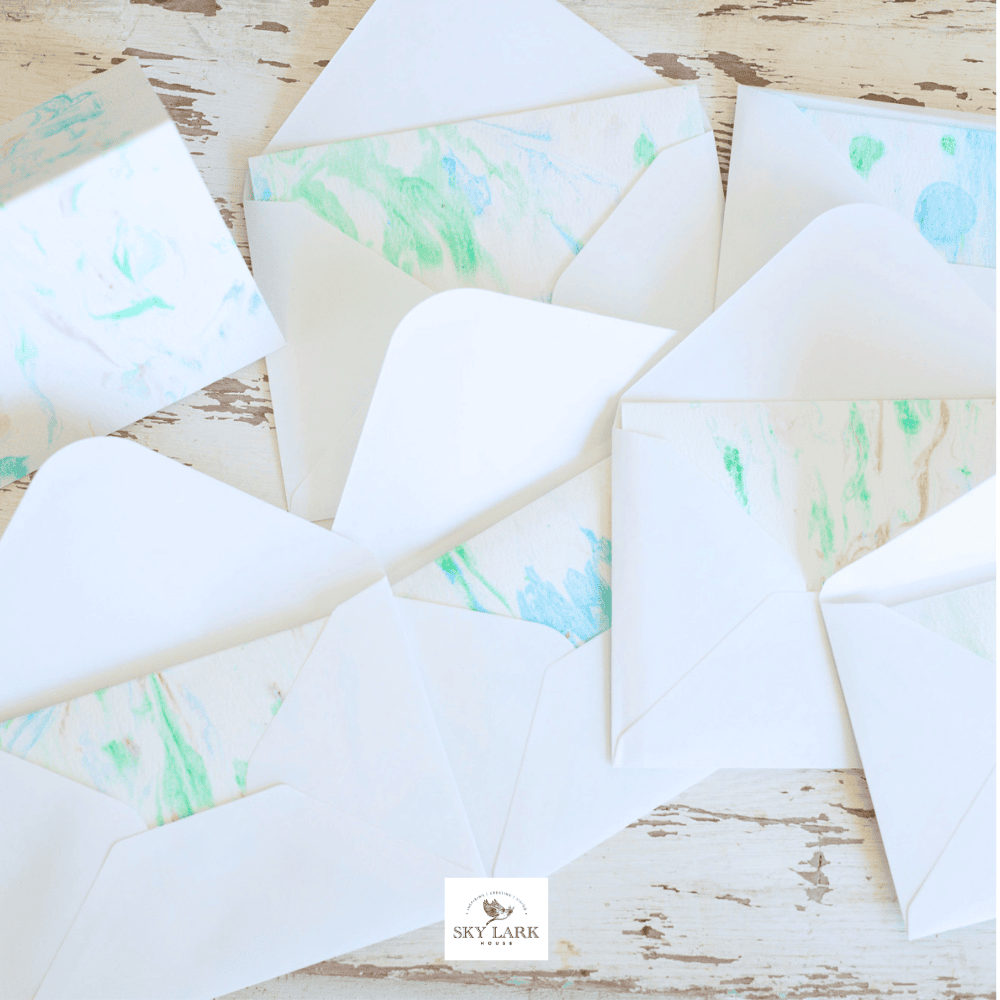 Several open white envelopes are arranged on a rustic wooden surface, each with a pastel marbled pattern lining in shades of blue, green, and pink. A small logo at the bottom reads "SKY LARK.