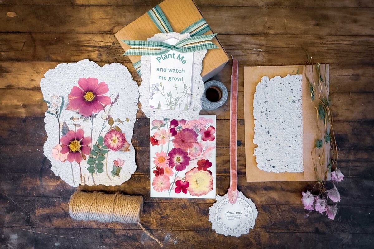 A flat lay of handmade paper with pressed flowers, a card reading "Plant Me and watch me grow!", twine, ribbon, dried flowers, a small ink bottle, and kraft paper on a rustic wooden surface.