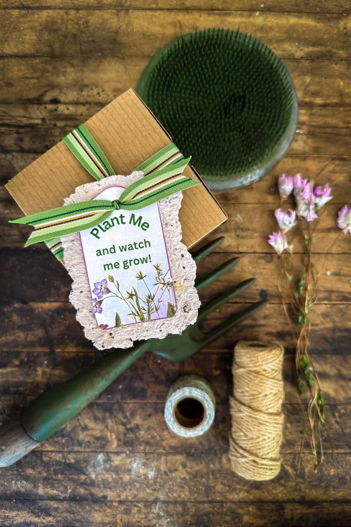 A gift box with a "Plant Me and watch me grow!" tag, green ribbon, and floral illustration sits on a rustic table with twine, dried flowers, a garden fork, and a green floral foam disk.