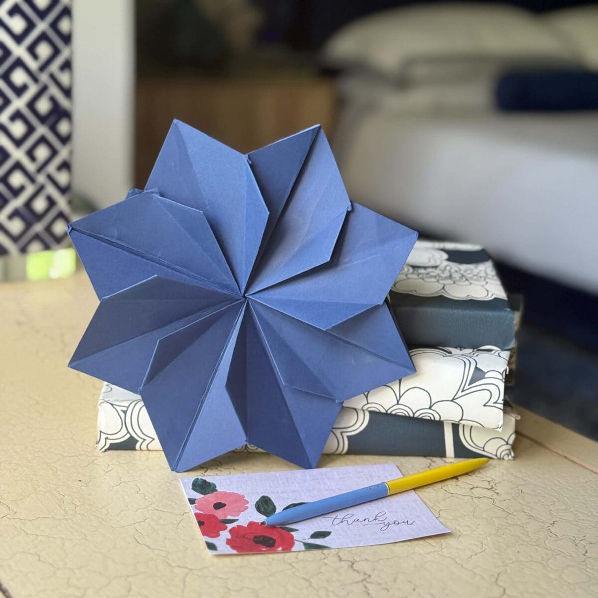 A large blue origami star sits on top of two stacked, patterned gift boxes. In front, there is a thank you card with floral design and a yellow pen on a light-colored table.