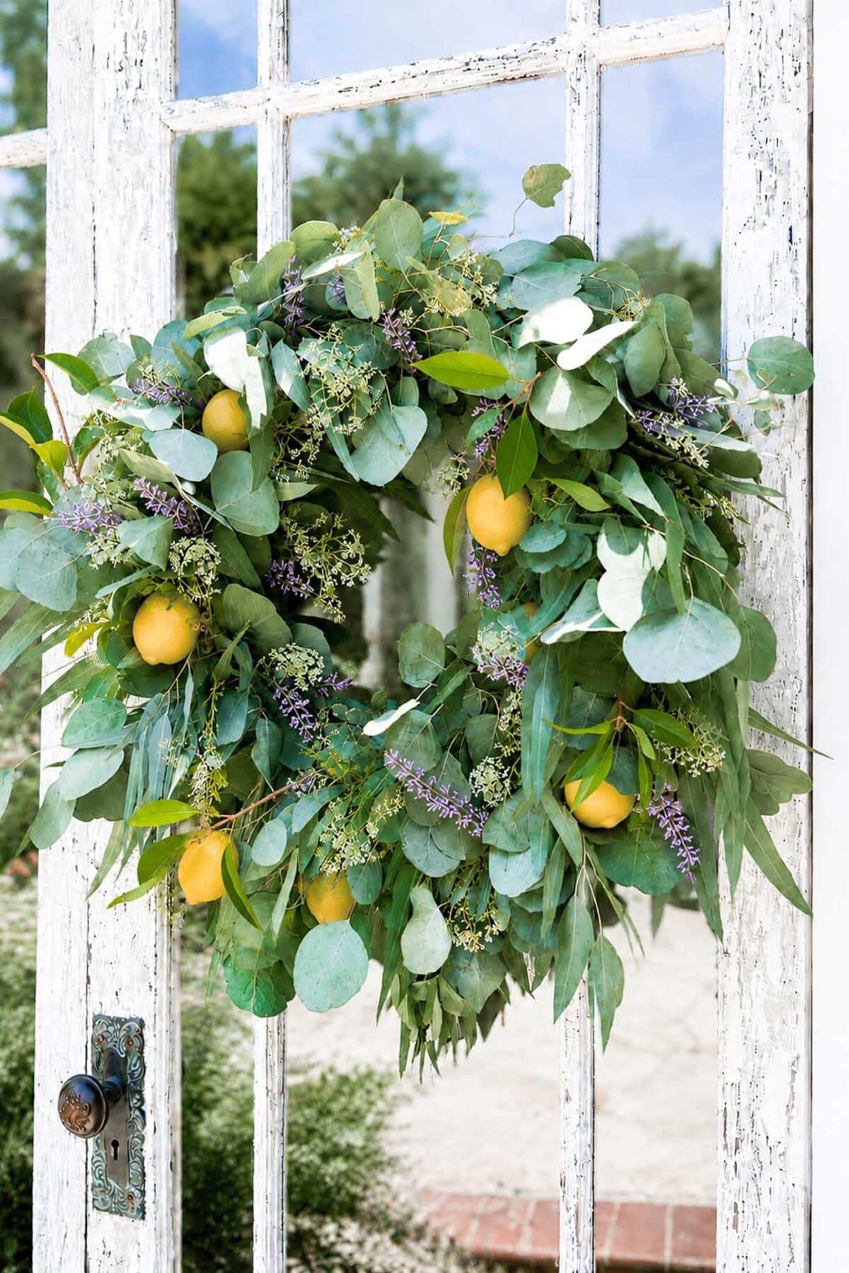A rustic wreath of eucalyptus leaves, lemons, and tiny purple flowers hangs on a weathered white wooden door with an antique doorknob.