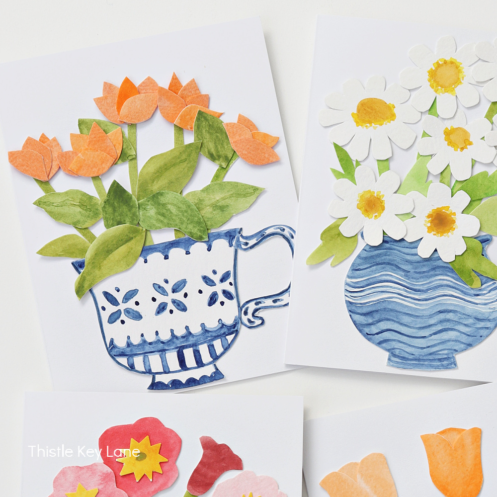 Three greeting cards featuring handmade paper flower designs: orange tulips in a blue teacup, white daisies with yellow centers in a blue vase, and a glimpse of colorful flowers on another card below.