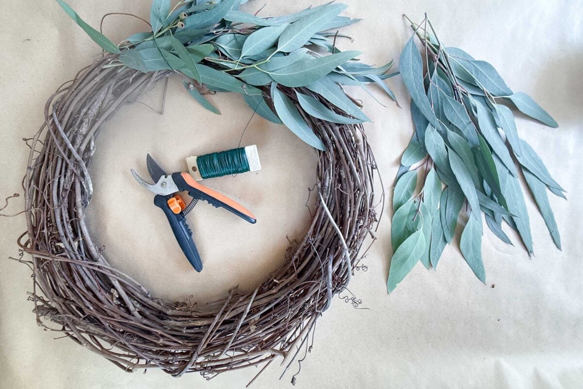 A grape vine wreath is laid out with eucalyptus branches, pruning shears, and floral wire for crafting.