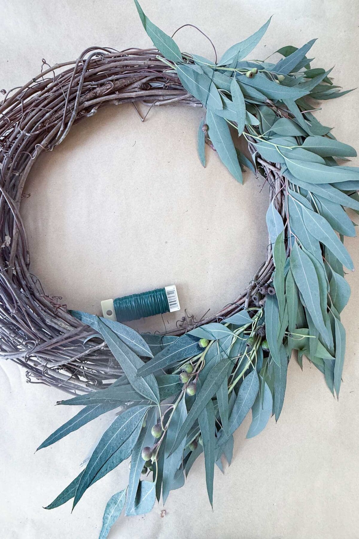 A grapevine wreath with eucalyptus branches and berries attached, and a spool of green floral wire.