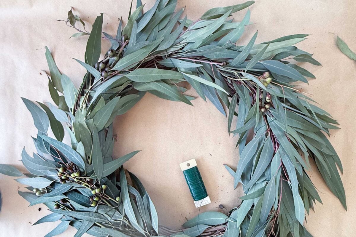 A eucalyptus wreath is being made on a brown surface. A spool of green floral wire rests in the center.