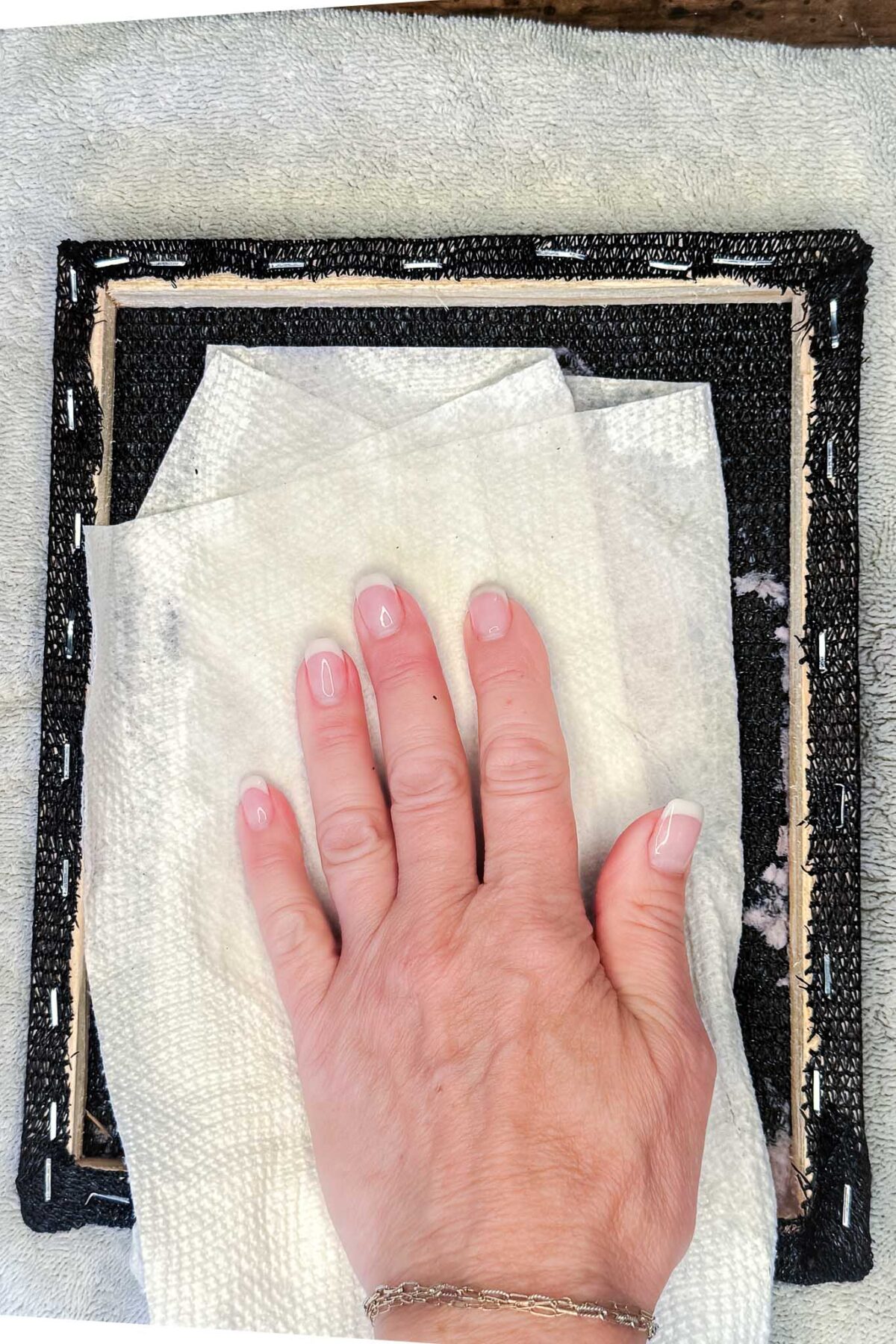 A hand with a silver bracelet wipes the back of a black-framed canvas using a white paper towel, perfect for creating DIY seed paper or handmade cards, on a light-colored textured surface.