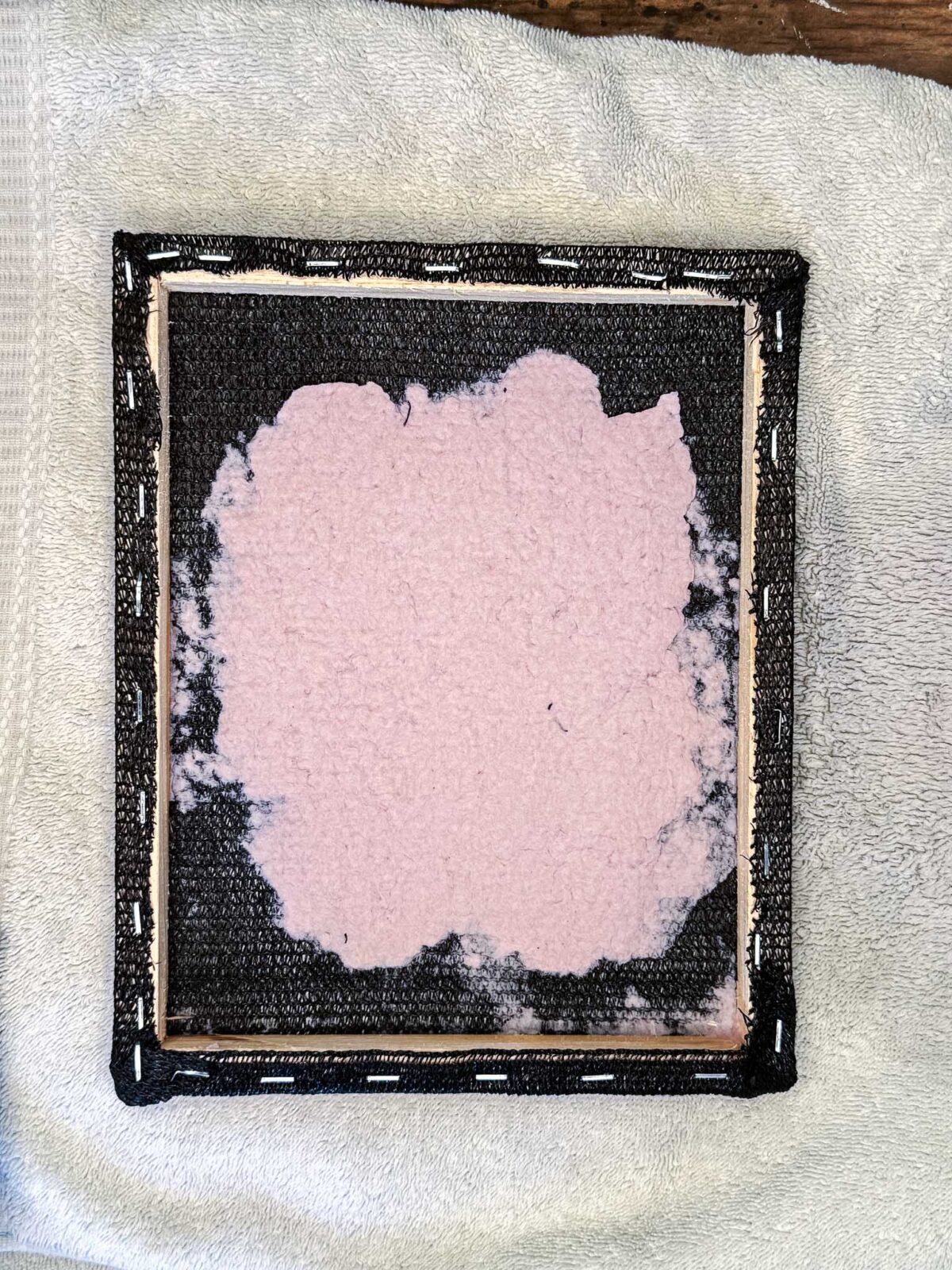 A square canvas with its edges stapled to a wooden frame lies on a white towel. The canvas surface has an irregular patch of light pink DIY seed paper pulp applied to the center, perfect for crafting handmade cards or unique stationery.