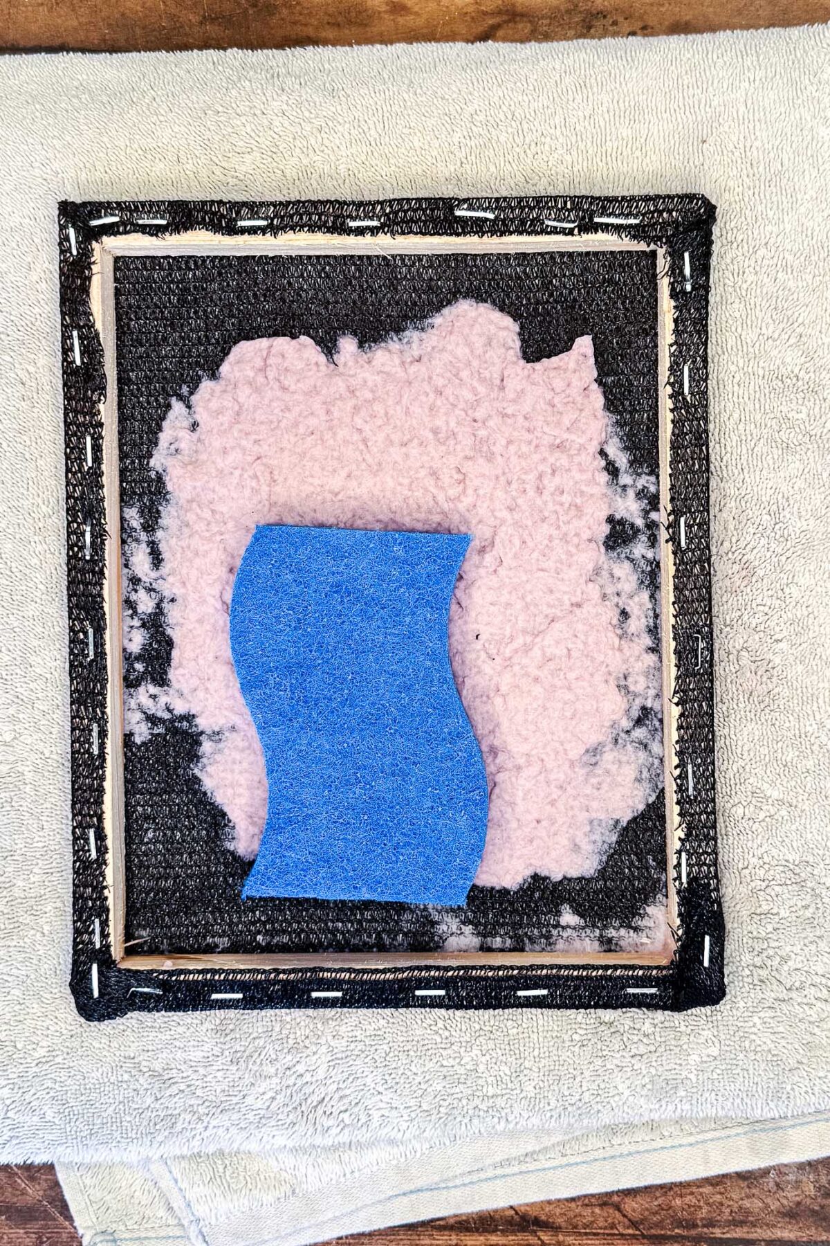 A blue sponge rests on pink and black foam inside a square wooden frame, placed on a light gray towel—perfect for crafting handmade cards or DIY seed paper as part of your creative stationery projects. The frame is stapled with black fabric around the edges.