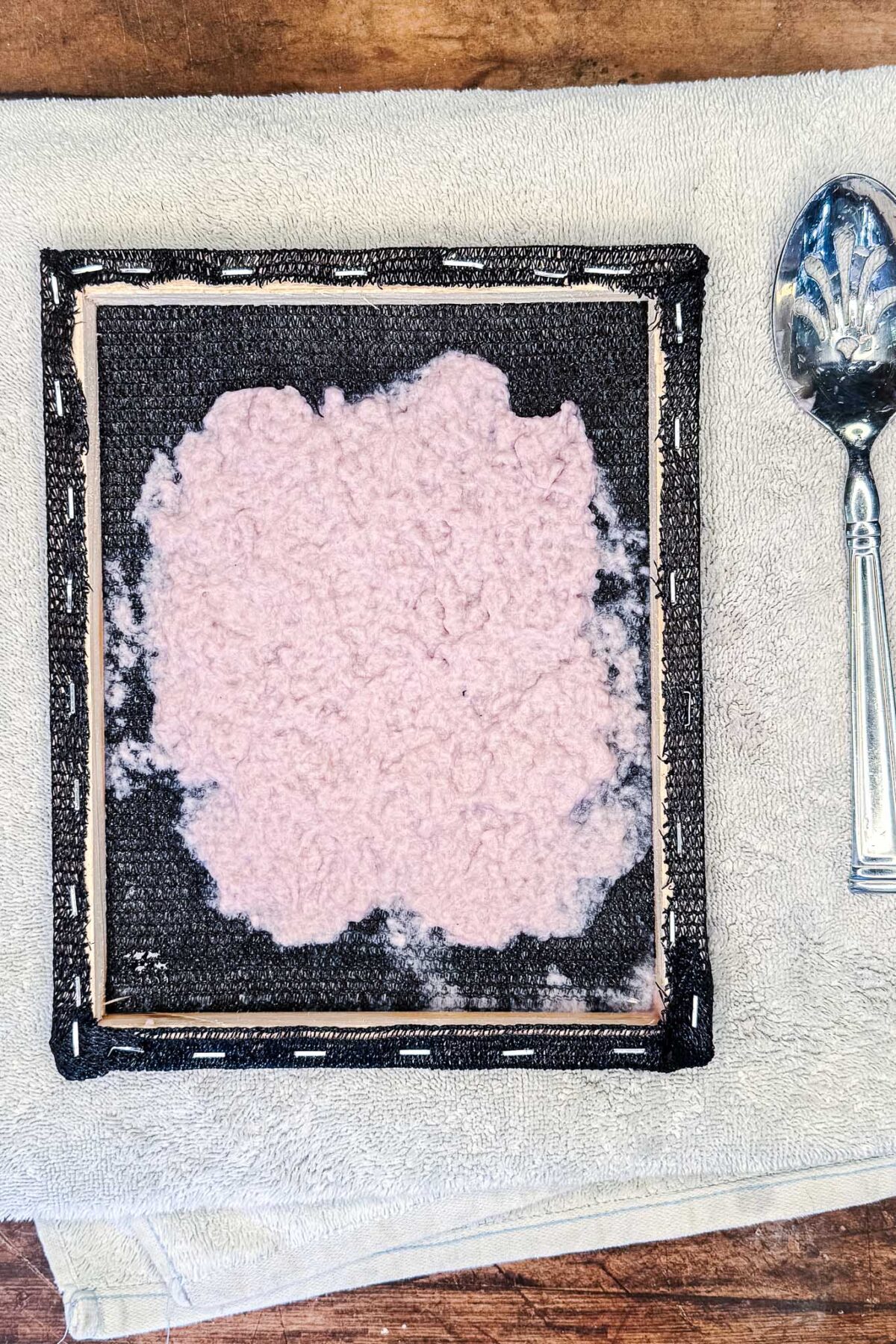 A square frame with a screen holds a sheet of wet, pale pink pulp—likely DIY seed paper for handmade cards or stationery—placed on a folded towel. A silver spoon with a decorative handle lies to the right on the towel.