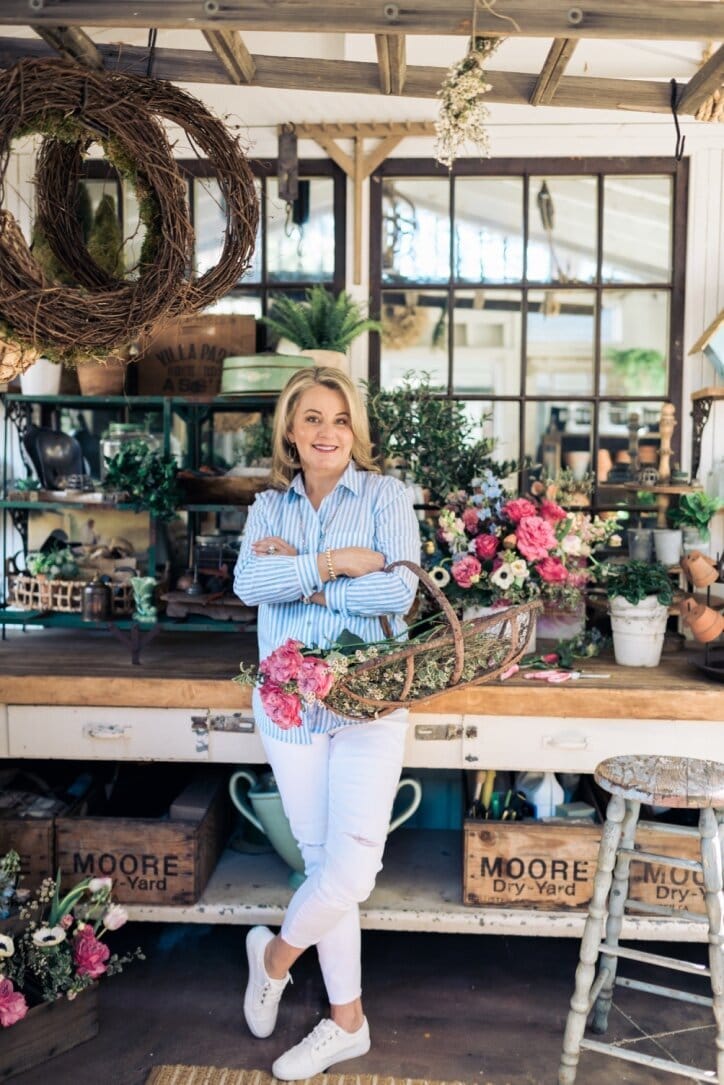A woman in a striped shirt and white pants stands in a flower shop, smiling and holding a basket of colorful flowers. The shop has wreaths, plants, and garden tools visible in the background.