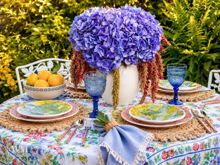 A round outdoor table with a large bouquet of purple hydrangeas in a white vase, blue glasses, colorful plates, woven placemats, a bowl of oranges, and a floral tablecloth. Lush greenery is in the background.