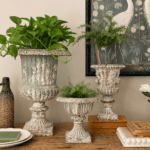 Three vintage-style urn planters with an aged patina, filled with green plants, sit on a wooden table in front of a crane artwork. Books, a woven vase, and stacked plates are also displayed in the arrangement.