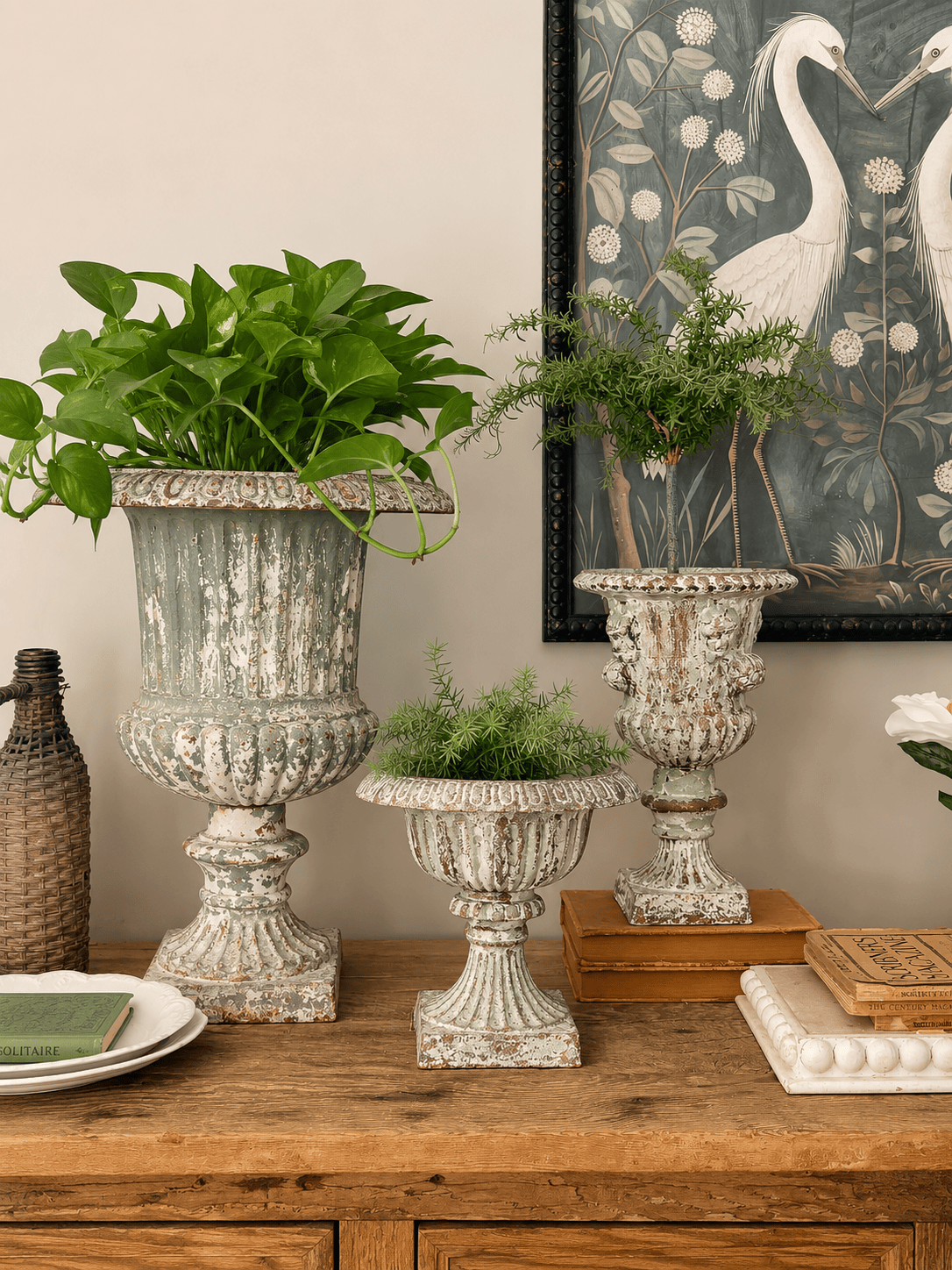 Three vintage-style urn planters with an aged patina, filled with green plants, sit on a wooden table in front of a crane artwork. Books, a woven vase, and stacked plates are also displayed in the arrangement.