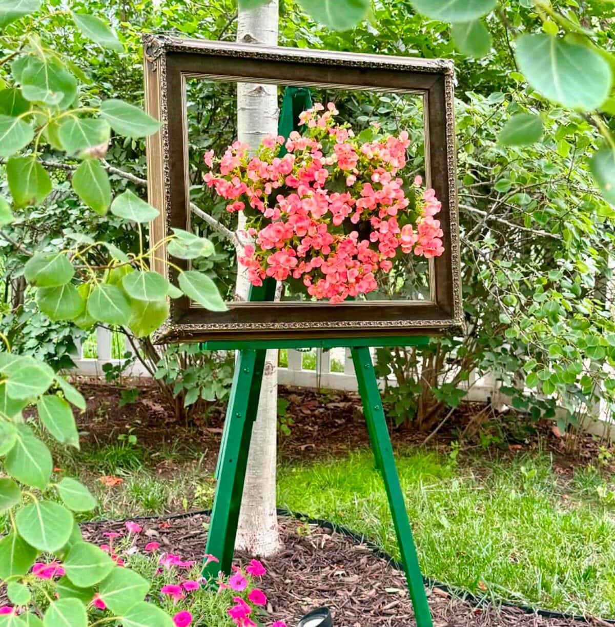 A vintage picture frame on a green easel stands outdoors, framing pink flowers and a tree trunk, creating the illusion of a living floral painting among green leaves and a garden background.