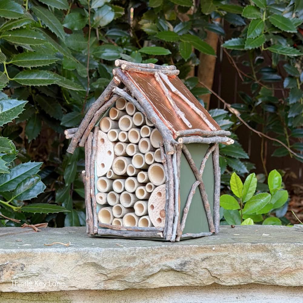 A small insect house made of sticks, bark, and bamboo tubes sits on a stone surface, surrounded by green foliage in the background.