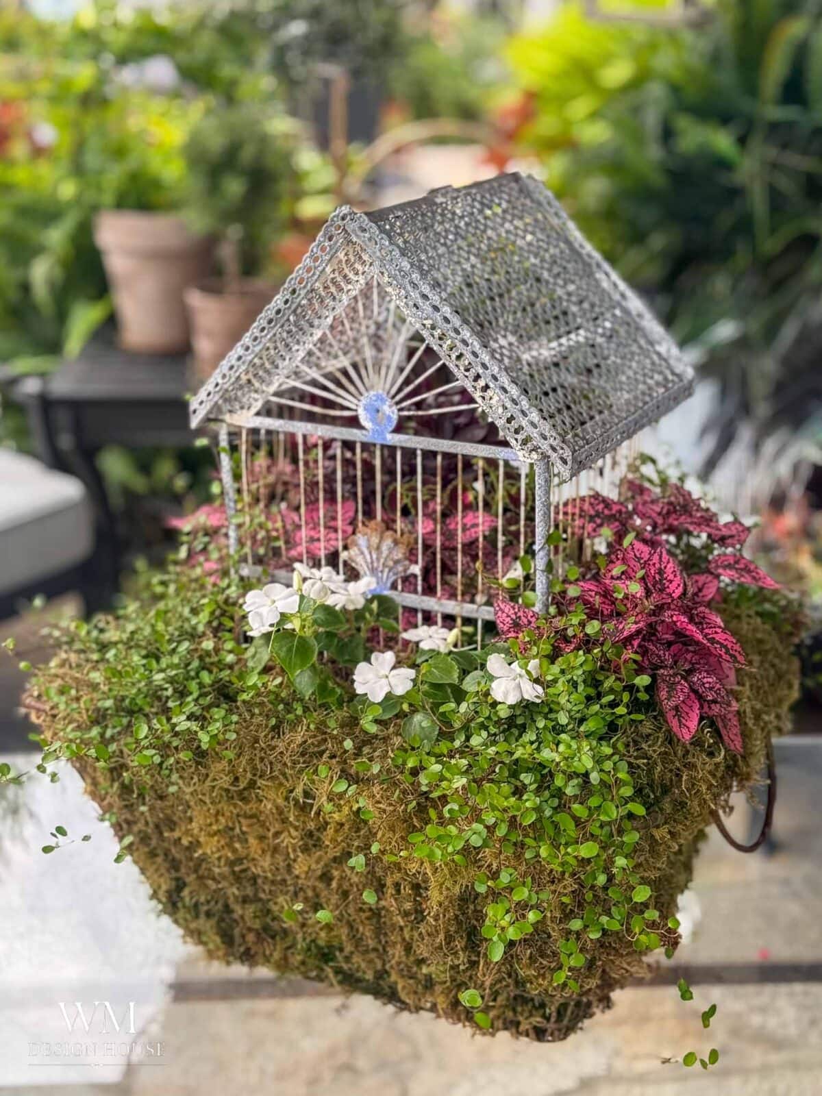 A decorative birdcage filled with lush green plants, white flowers, and pink foliage sits atop a moss-covered base, surrounded by potted plants in a garden setting.