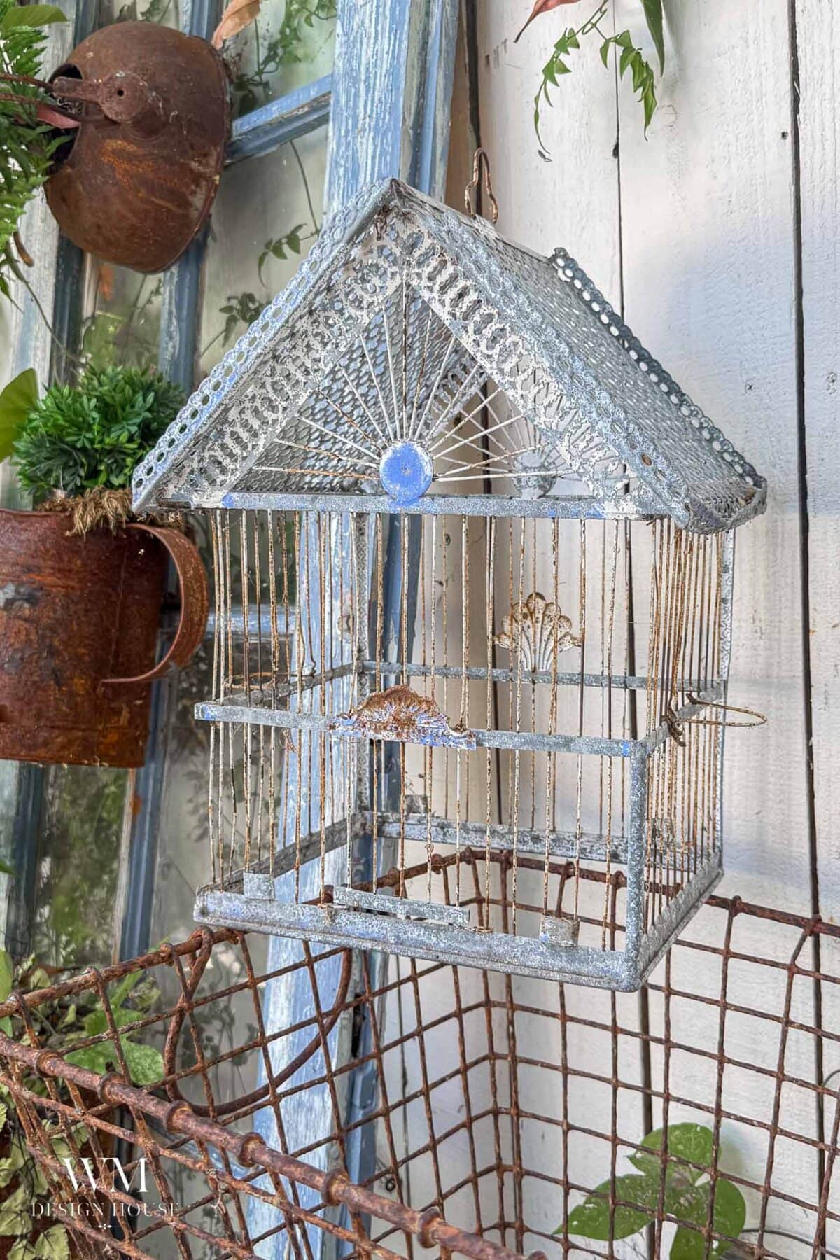 A decorative, vintage-style empty birdcage planter with a pointed roof hangs beside a rusty watering can and a wire basket on a weathered wooden wall with plants nearby.