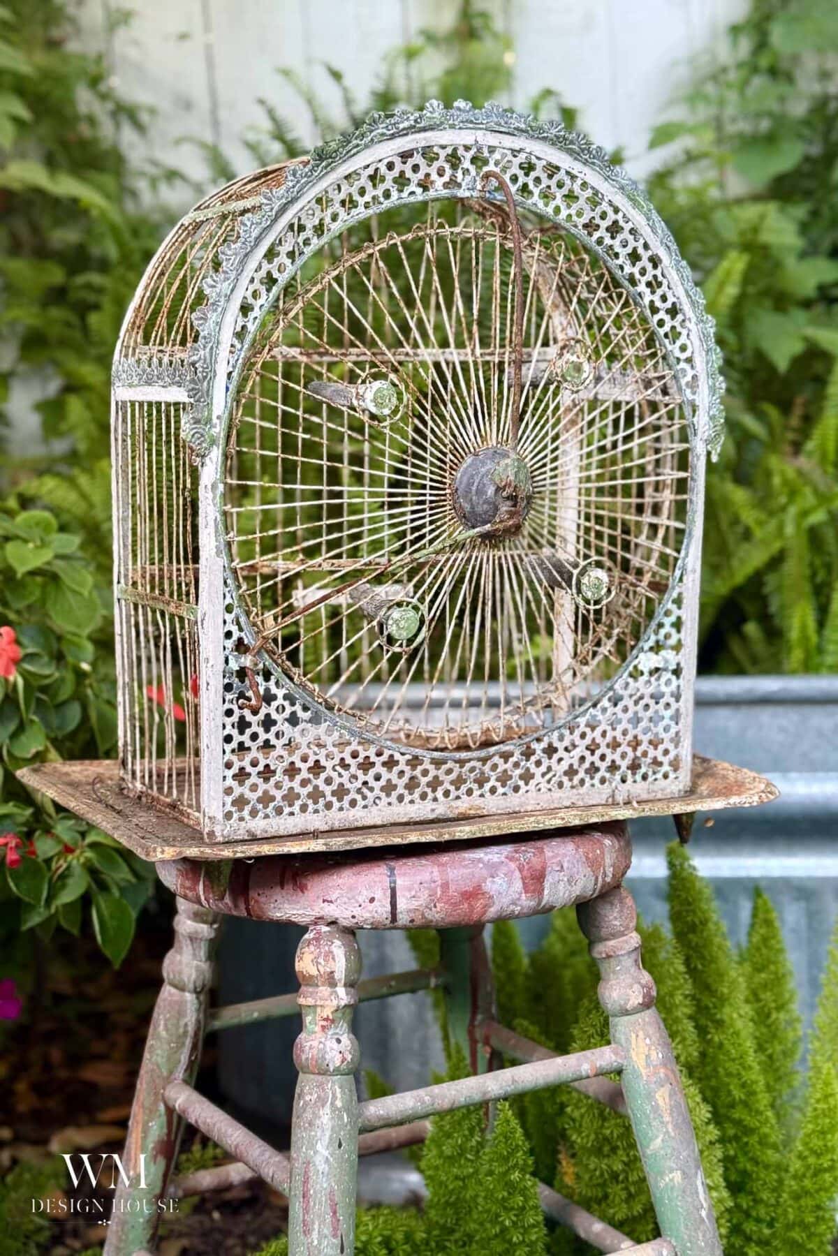A vintage, ornate metal birdcage planter with a rounded top sits on a weathered, multicolored wooden stool, surrounded by lush green plants and flowers in an outdoor garden setting.