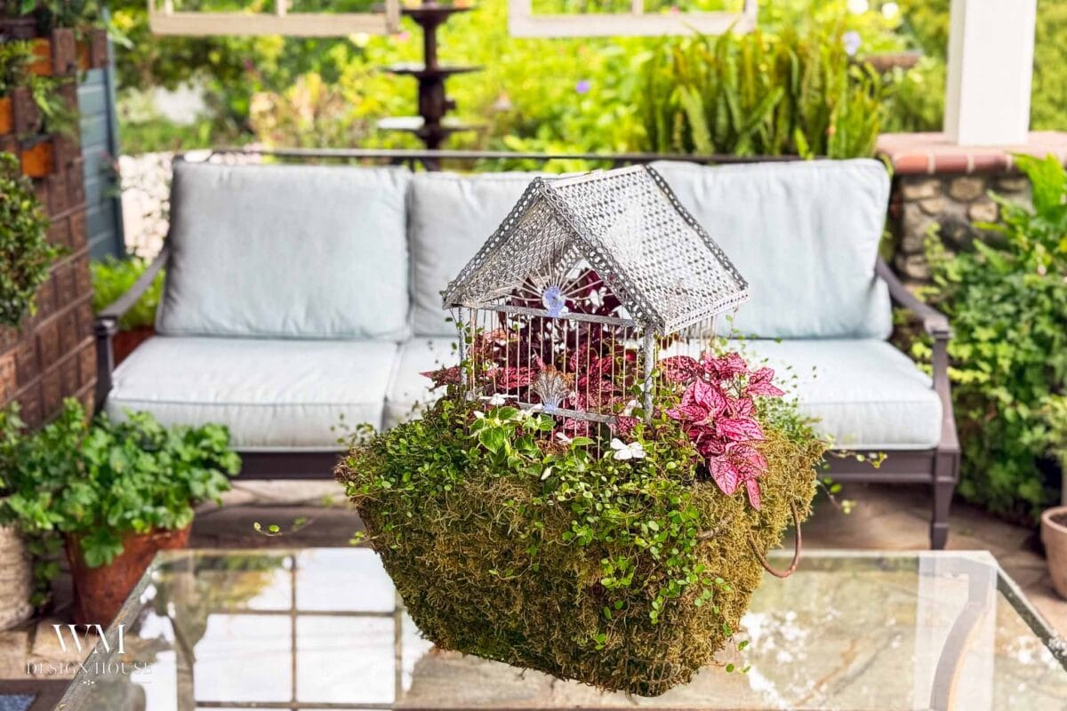 A charming Birdcage Planter filled with pink and green foliage sits on a glass table in front of a white cushioned outdoor sofa, surrounded by lush greenery.