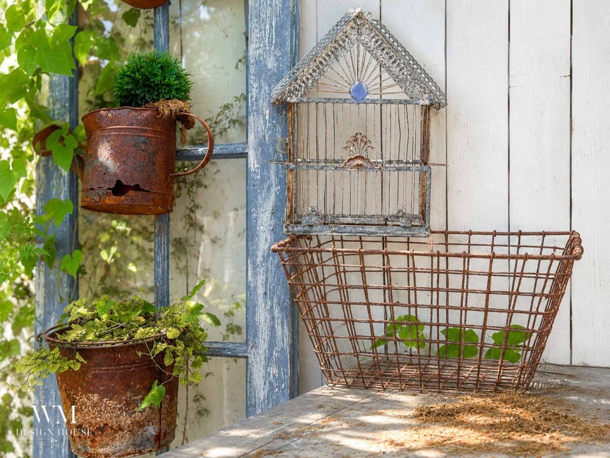 Rusty pots with plants hang on a window frame next to a white wall with a wire birdhouse decor. Nearby, a charming Birdcage Planter adds vintage flair, while a wire basket with a few green leaves sits on a weathered wooden surface. Sunlight filters through leaves.
