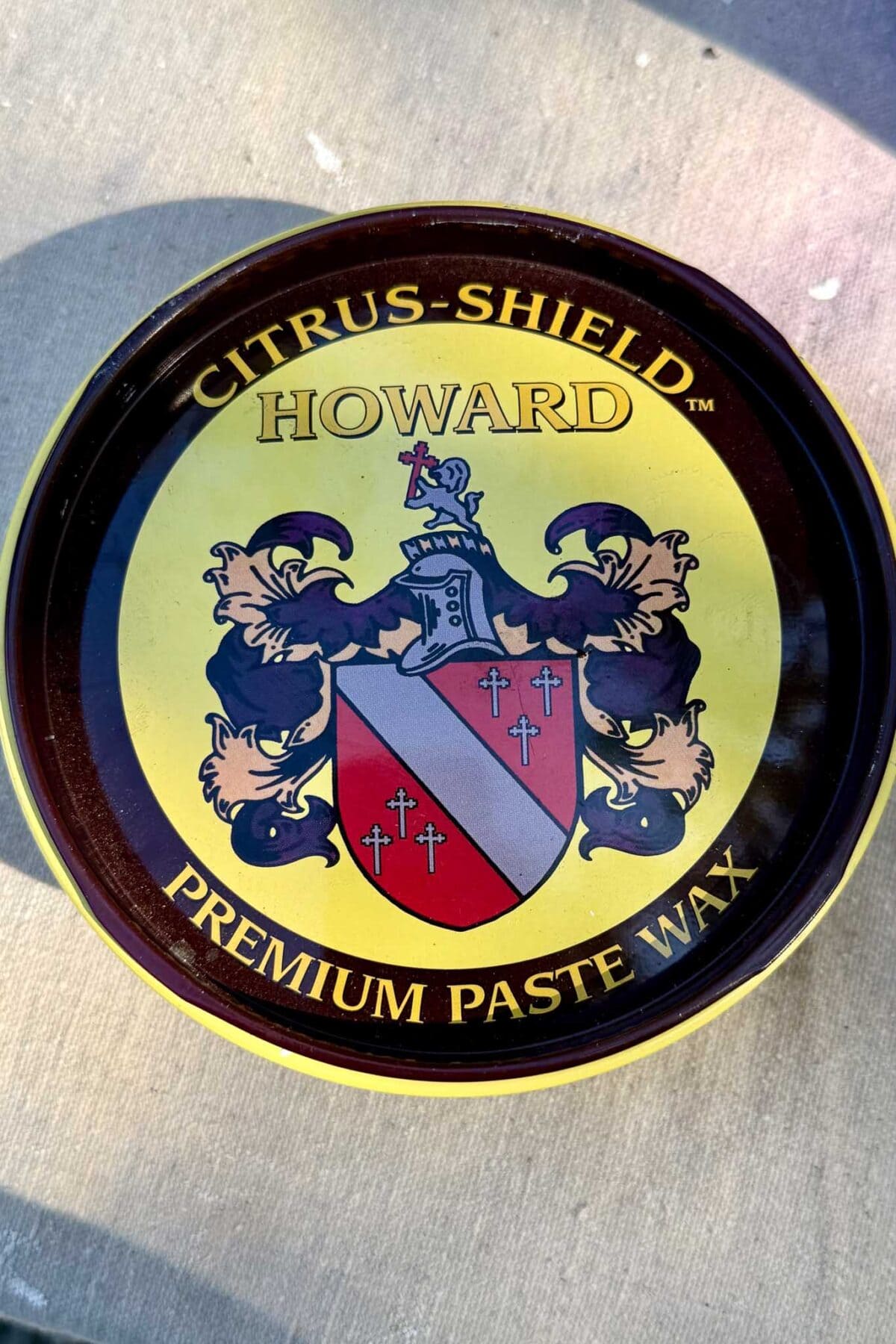 A round tin labeled “CITRUS-SHIELD HOWARD PREMIUM PASTE WAX,” featuring a crest with a knight’s helmet, shield, and decorative elements on a yellow background—perfect for enhancing an aged patina or reviving rusted urns with a rich, lustrous patina finish.