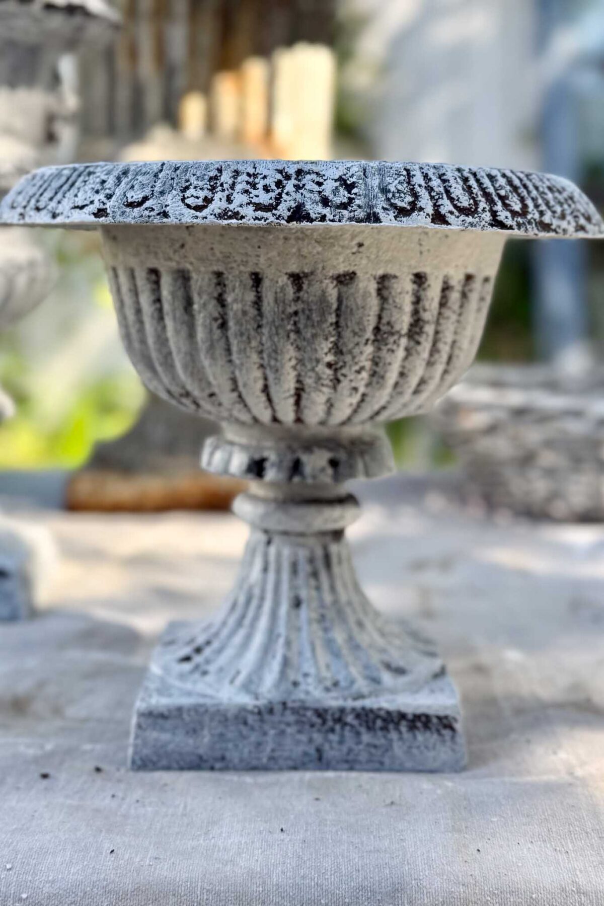 A weathered, ornate stone urn with a fluted design stands on a square base atop a light-colored surface, showcasing how to give an aged patina finish for timeless character, set against a blurred natural background.
