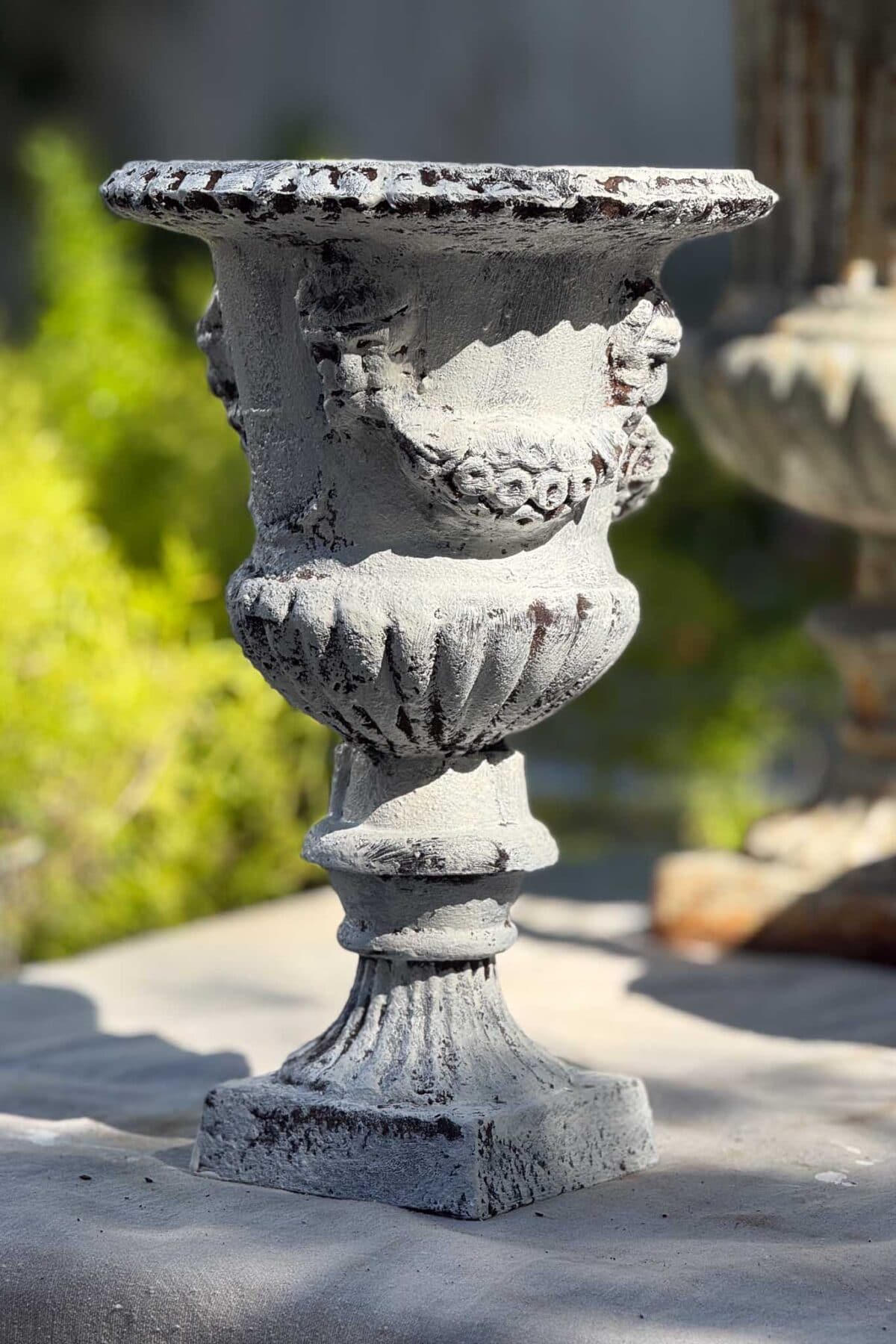 A weathered, ornate stone urn with carved details stands on a sunlit surface outdoors, surrounded by blurred greenery in the background.