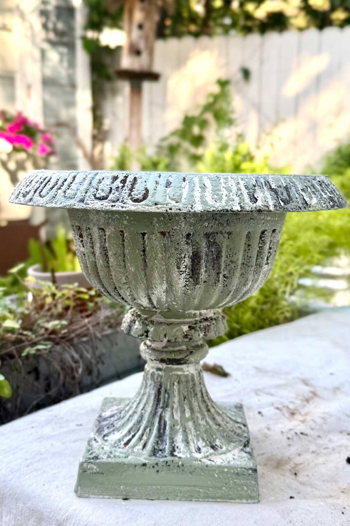 A weathered, ornate metal urn with a fluted design sits on a table outdoors, surrounded by greenery and flowers, with a white fence in the background.