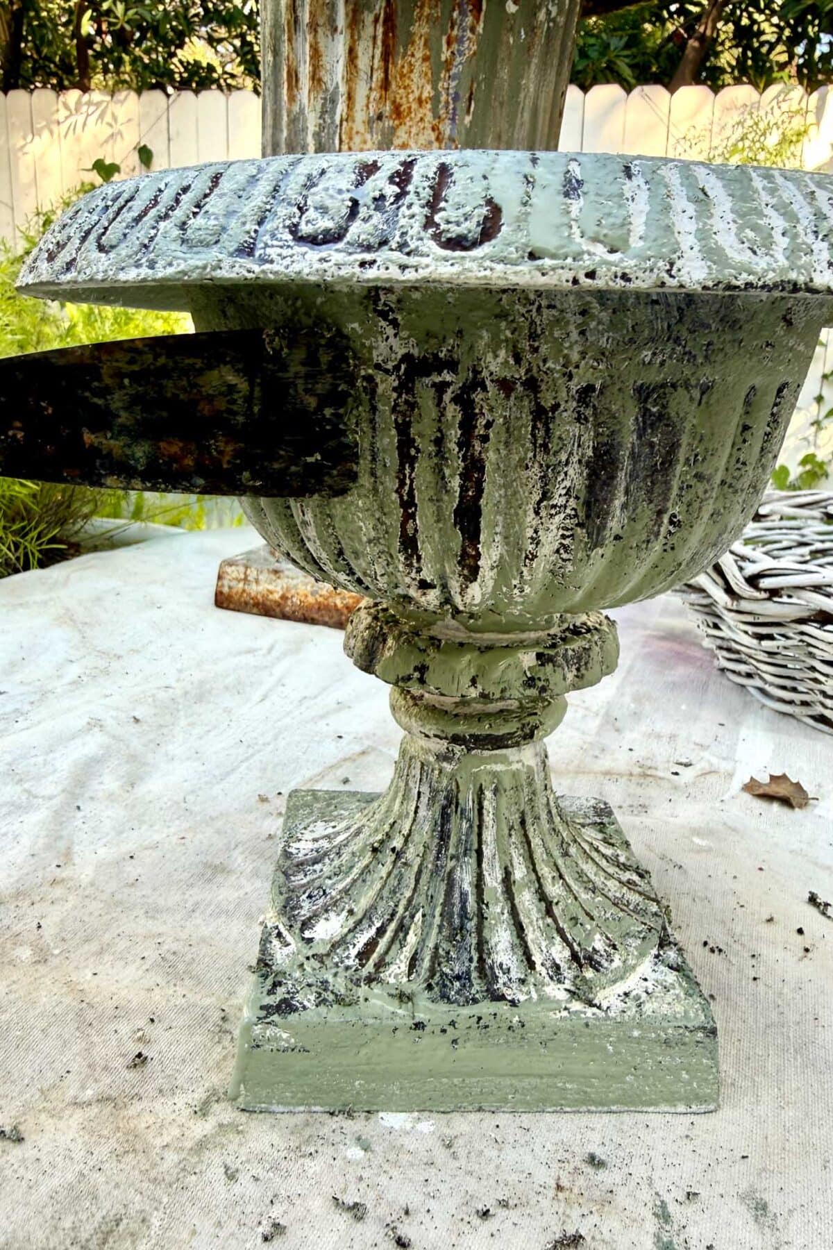 A close-up of a weathered green urn with peeling paint. A rusty tool scrapes the side, and a white fence and greenery are visible in the blurry background.