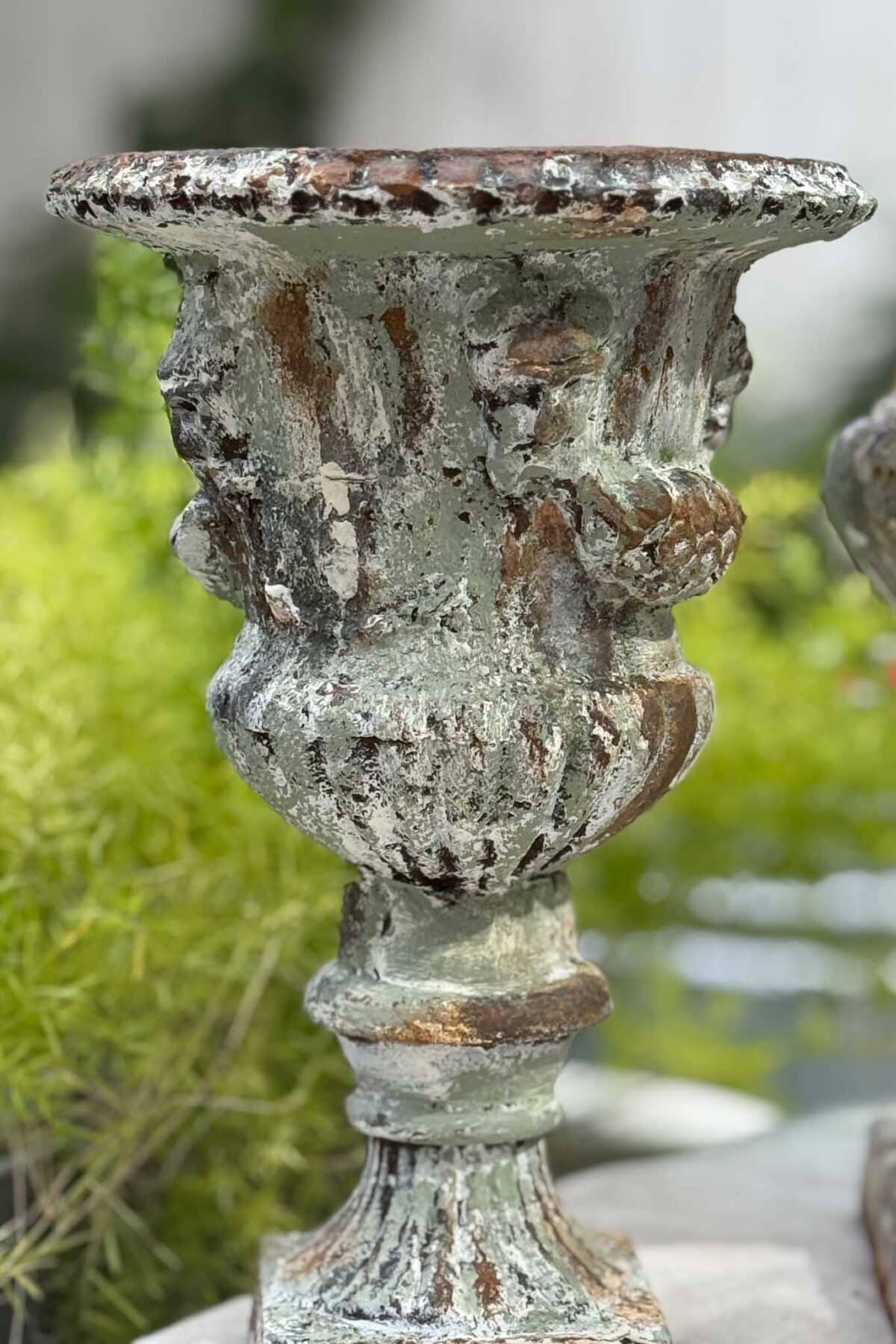 A weathered, ornate stone urn with faded green and brown patina stands outdoors, surrounded by blurred green foliage in the background.