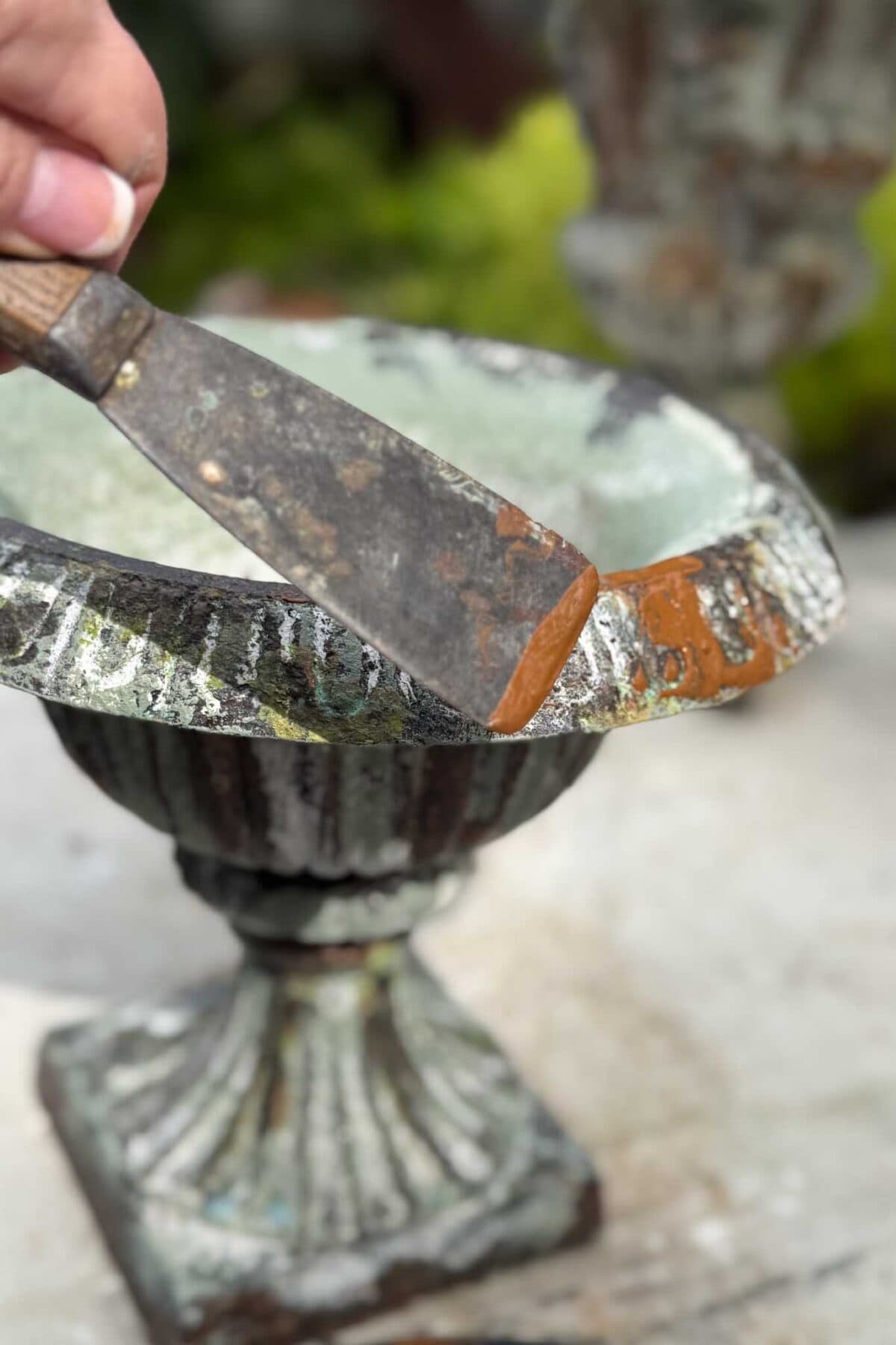 A hand scrapes rust off the rim of a weathered, ornate metal planter with a putty knife.