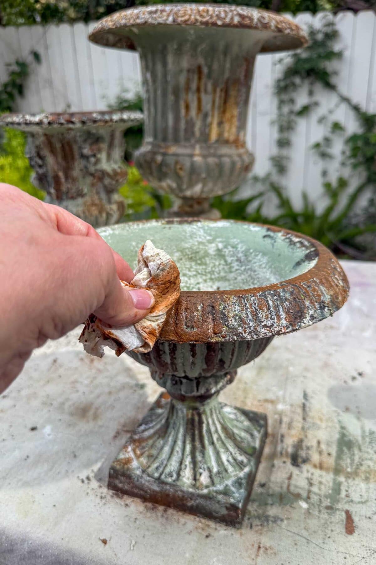 A hand uses a cloth to clean the rim of a weathered, rusty metal urn outdoors. Another similar urn and a white fence with greenery are visible in the background.
