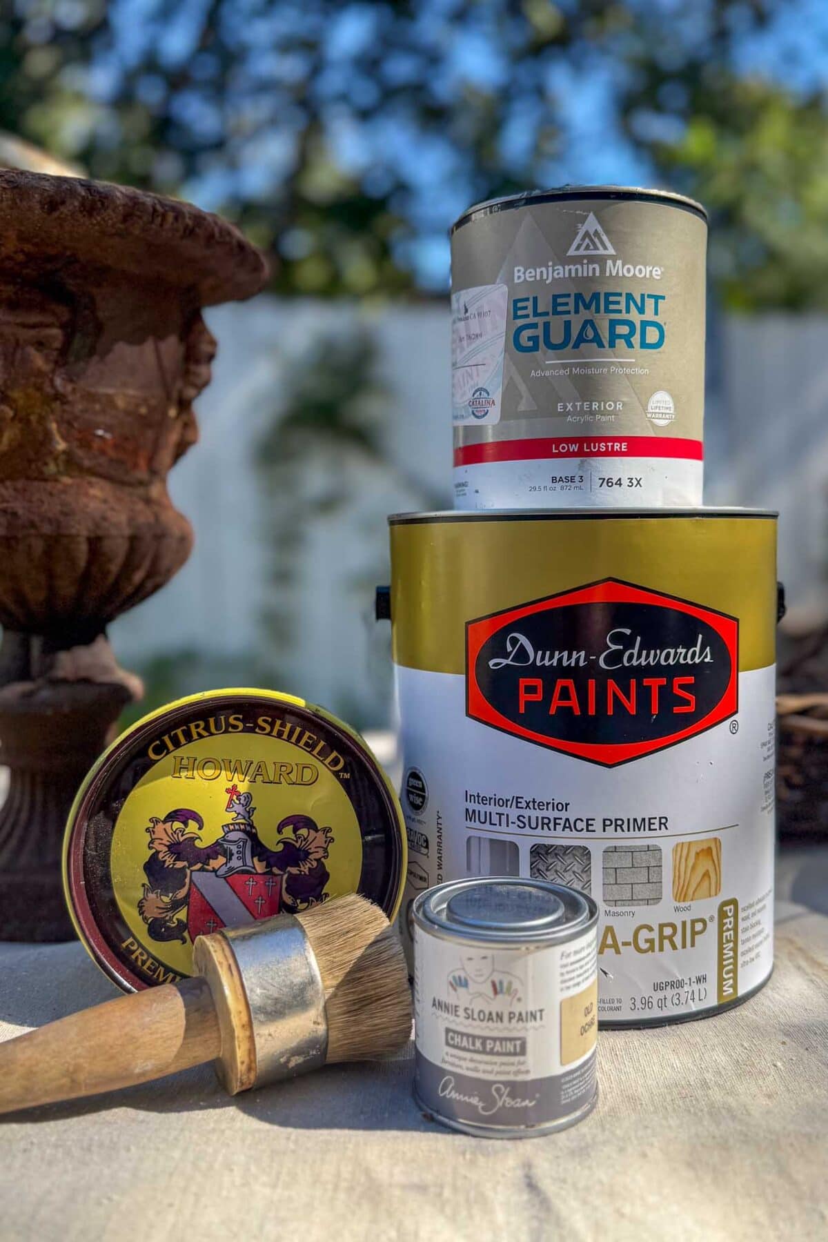 A close-up of various paint cans, primer, a wax tin, and two paintbrushes on a table outdoors, with blurred plants and a fence in the background.