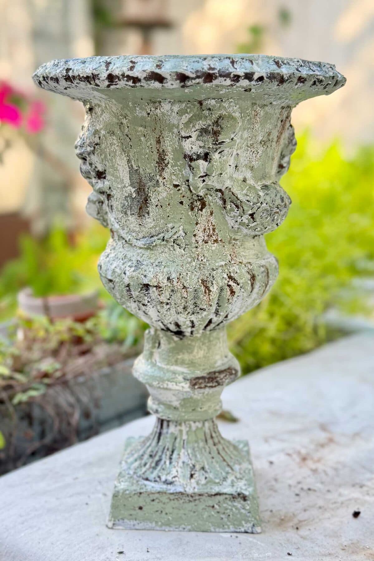 A weathered, greenish stone urn with ornate details stands on a white surface outdoors, with blurred greenery and flowers in the background.