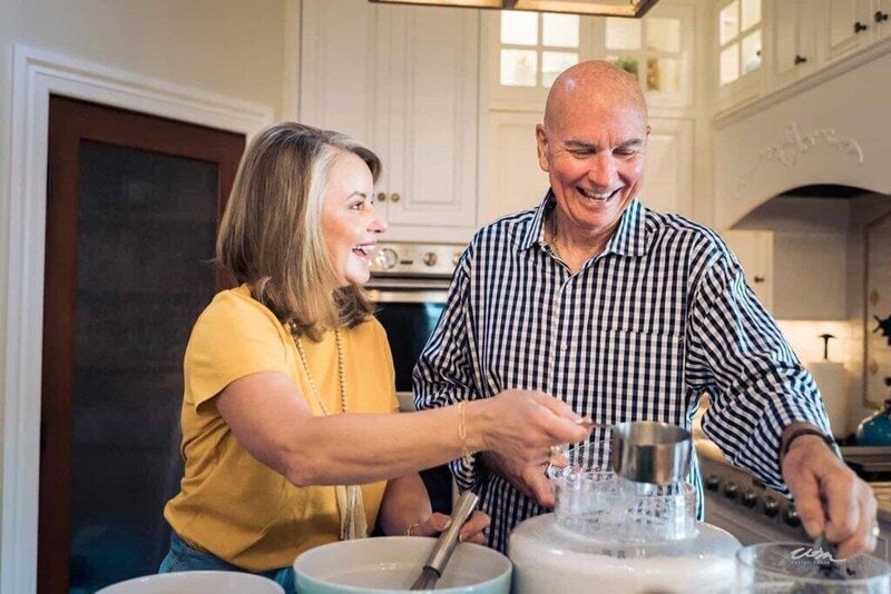 A smiling man and woman prepare food together in a bright kitchen, using a mixer and measuring ingredients.