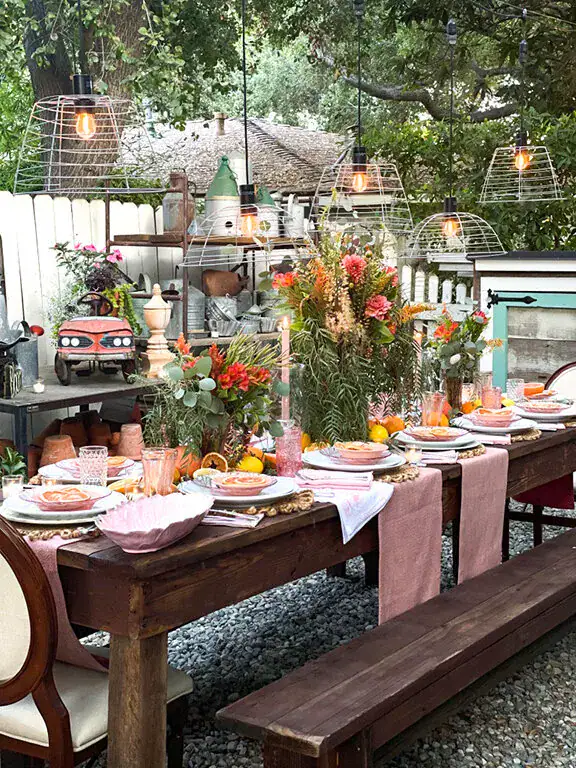 Outdoor dining table set for a meal with pink table runners, floral centerpieces, plates, glassware, and hanging lights, surrounded by garden decor and greenery.