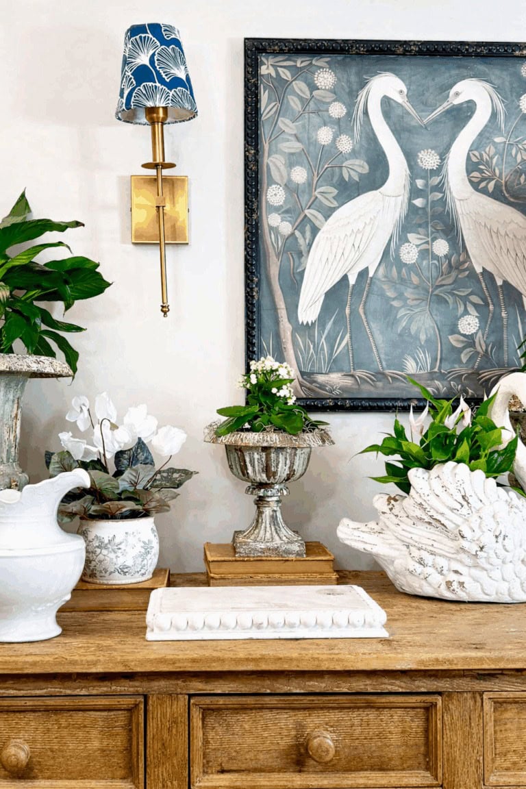 A wooden dresser displays potted plants, a white ceramic pitcher, and a swan planter. Above, a wall lamp with a blue shade and a framed artwork of two white herons are visible.