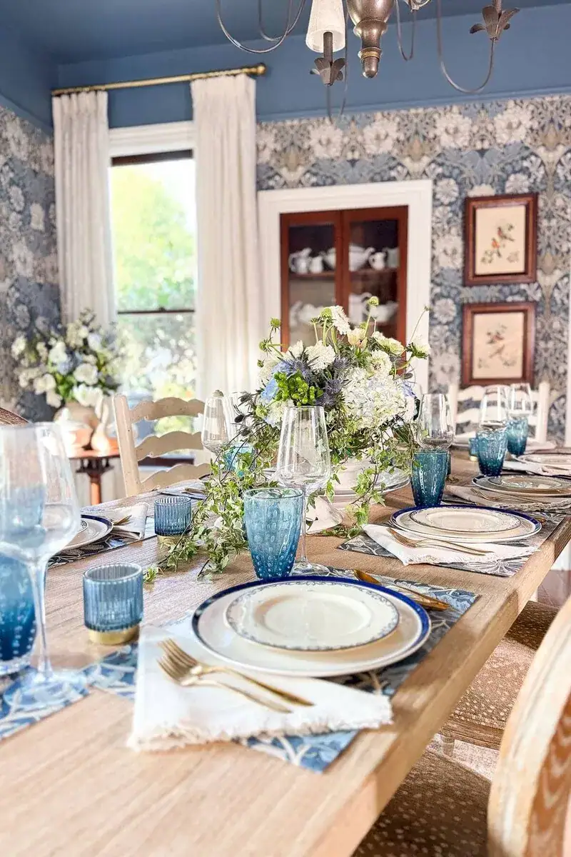 Elegant dining table set with blue glassware, white plates, gold cutlery, and floral centerpiece. Blue and white wallpaper, framed art, and light-filled window create a cozy, inviting atmosphere.