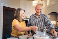 Older woman and man smiling and washing dishes together in a bright, modern kitchen. The woman wears a yellow shirt, and the man wears a checked shirt. They appear to be enjoying the chore.