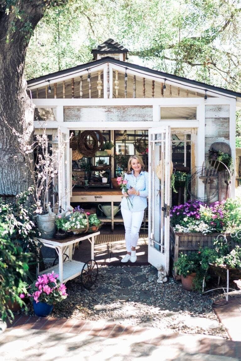 A woman stands smiling in the doorway of a sunlit garden shed, surrounded by blooming flowers, potted plants, and rustic garden decor.