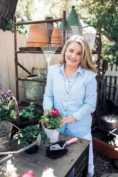 A woman in a blue striped shirt smiles while planting flowers at a garden table, surrounded by pots, tools, and plants in a sunlit outdoor setting.