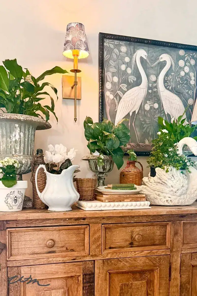 A wooden sideboard with potted plants, white ceramic pitchers, stacked books, and a framed artwork of two white birds beneath a wall sconce with a patterned shade.