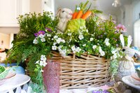 Wicker basket filled with greenery, white flowers, a small plush bunny, and fake carrots. Another bunny figure is next to the basket, creating a festive spring or Easter scene.