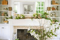 A bright living room with a white fireplace, floral arrangements on the mantel, and shelves filled with books, plants, and decorative items. A large mirror hangs above the mantel, reflecting a window.