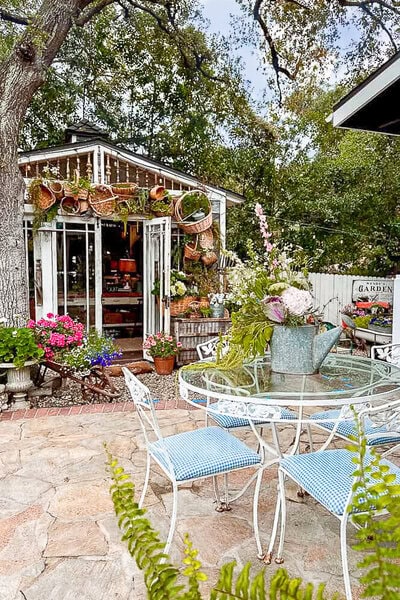 A charming garden patio with a glass table and blue-checkered chairs, surrounded by flowers, plants, and a shed decorated with hanging baskets and garden tools.