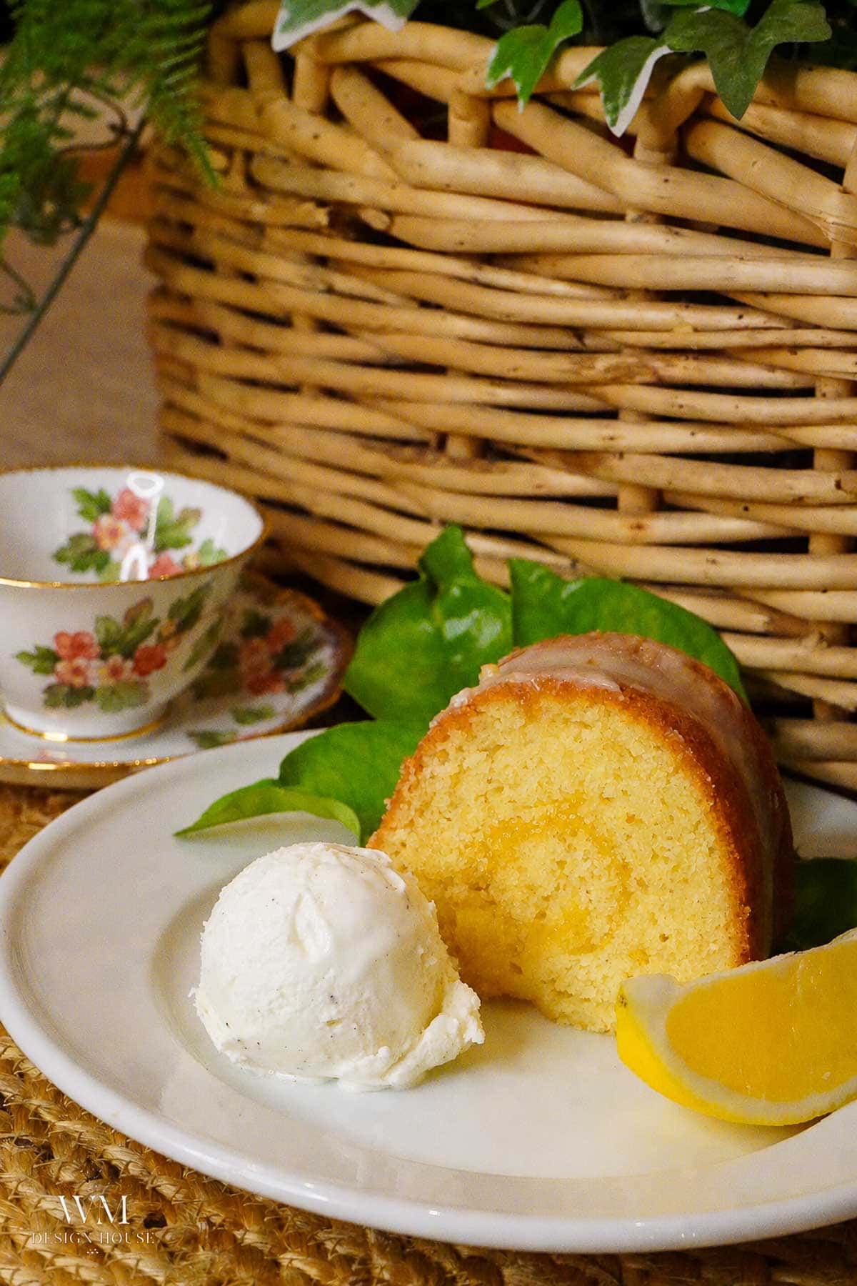 A slice of yellow bundt cake with a glaze, served with a scoop of vanilla ice cream and a lemon wedge on a white plate. A tea cup and a woven basket are in the background.